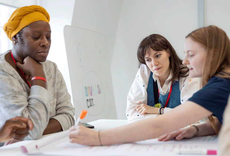 Image showing three students sitting at a table while one of them writes on a large sheet of paper with an orange marker pen