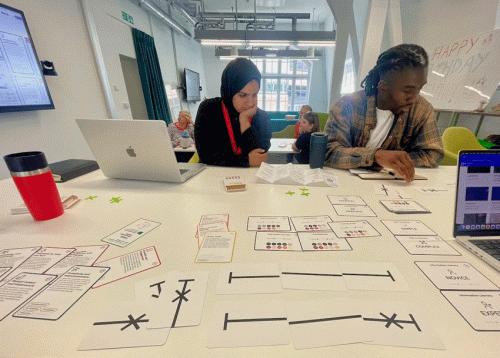 Diverse CDT students Sabah and Isaac are shows studying a table full of cards containing text and graphics as part of a data visualization challenge